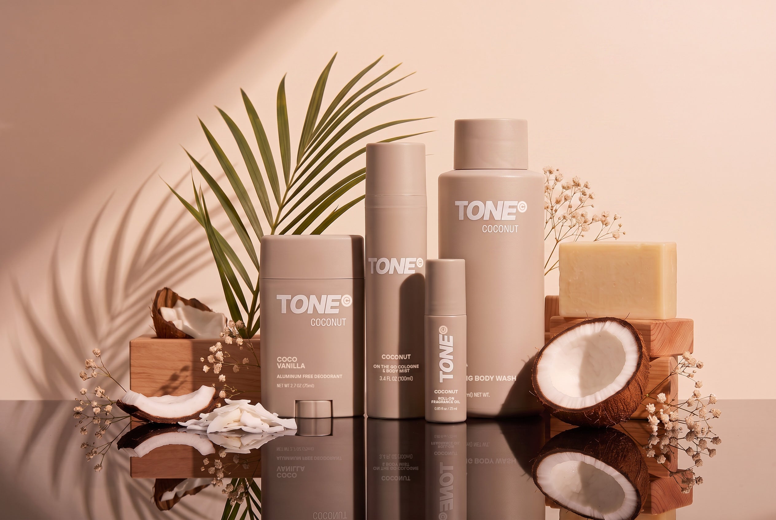 A set of TONE Coconut body care products, including a shower gel bottle, body wash bottle, and deodorant stick, styled with natural props like coconuts, a soap bar, and a palm leaf on a reflective surface against a warm beige background.