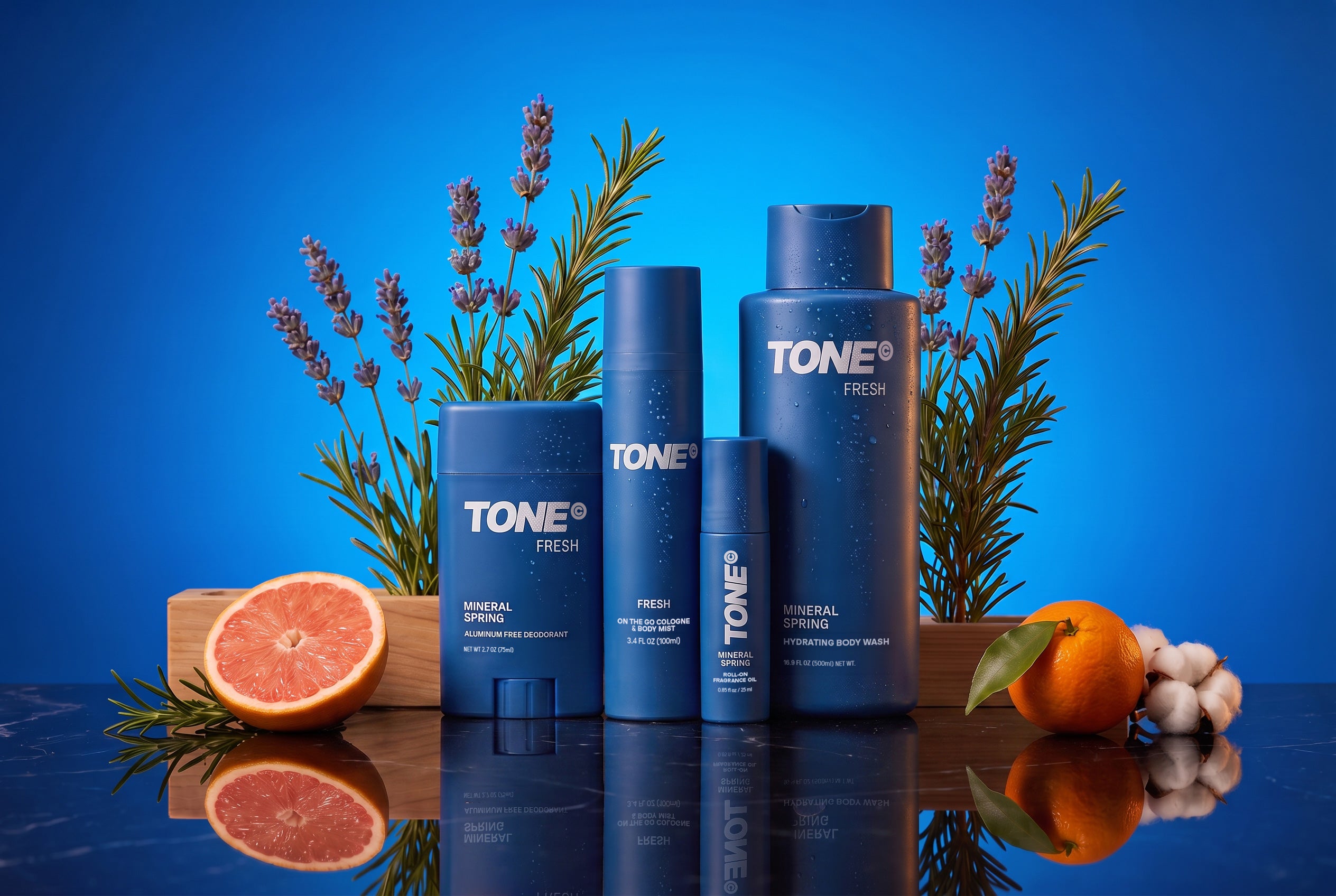 A set of blue TONE Fresh skincare products, including toner, spray, and body wash bottles, arranged on a reflective dark surface with grapefruit, orange, garlic, and lavender sprigs against a bright blue background.